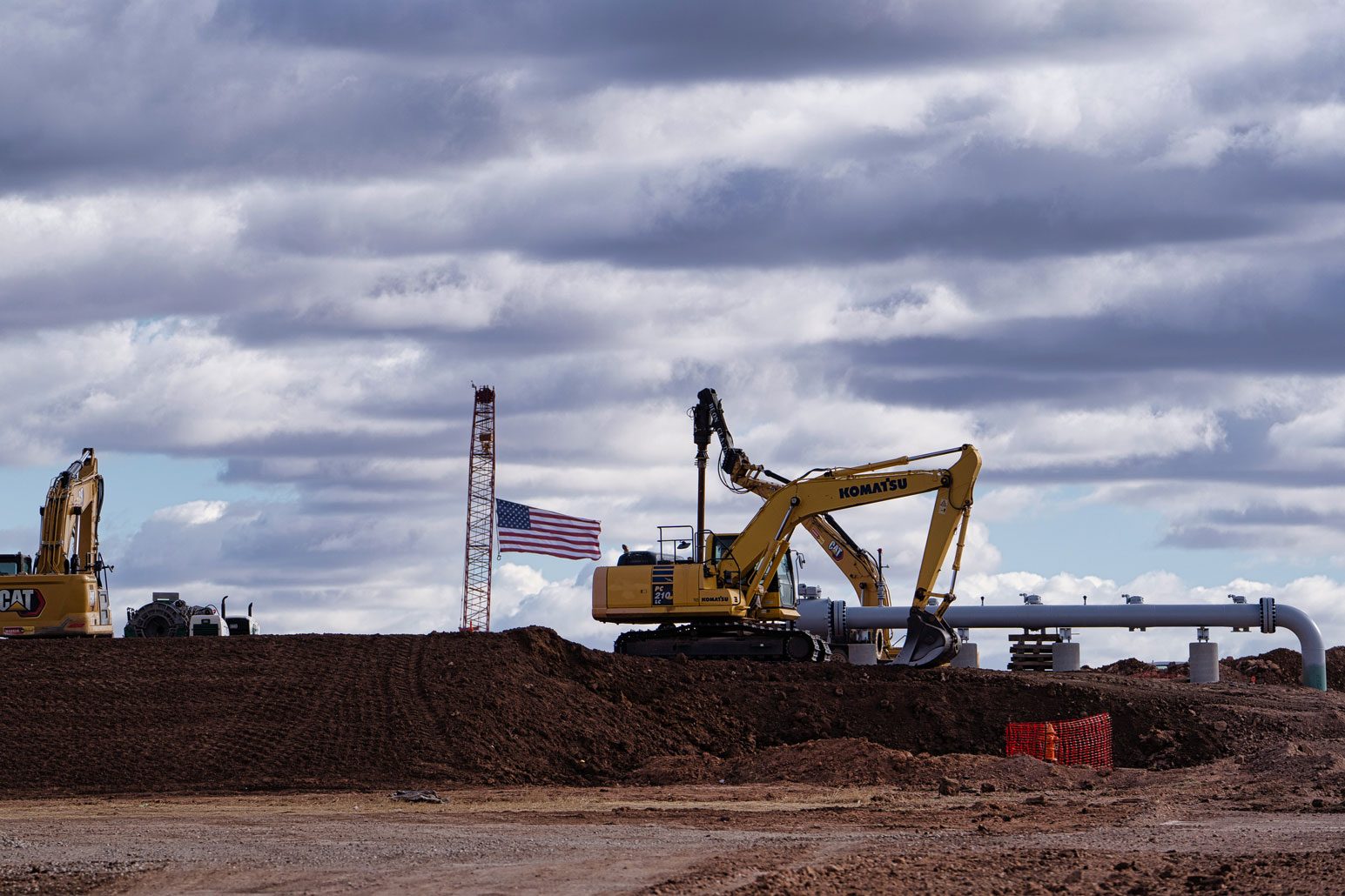 An excavator lowers a pipeline section into a trench beside an American flag on the Project Matador site